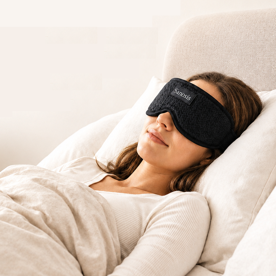 A woman lying in bed with her eyes closed, wearing a black textured sleep mask with the brand name 'Sanosis' visible on the front. The scene is softly lit with neutral tones, featuring white pillows and beige bedding.