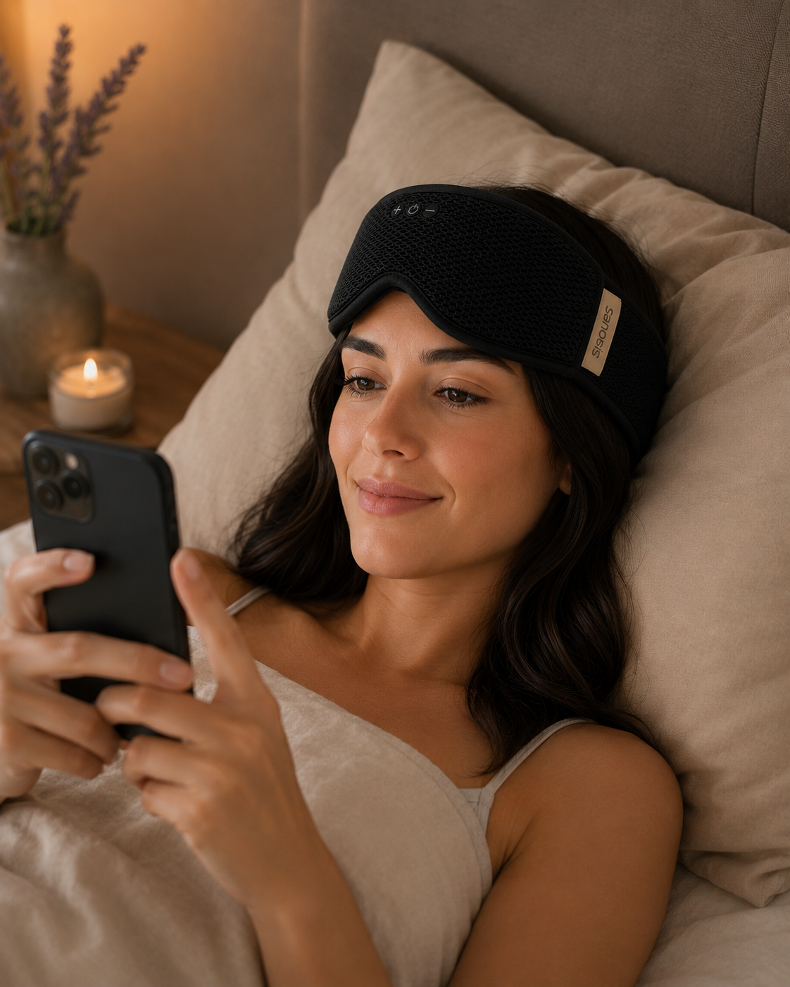 Woman in bed using a phone with a black sleep mask on, in a cozy bedroom setting.