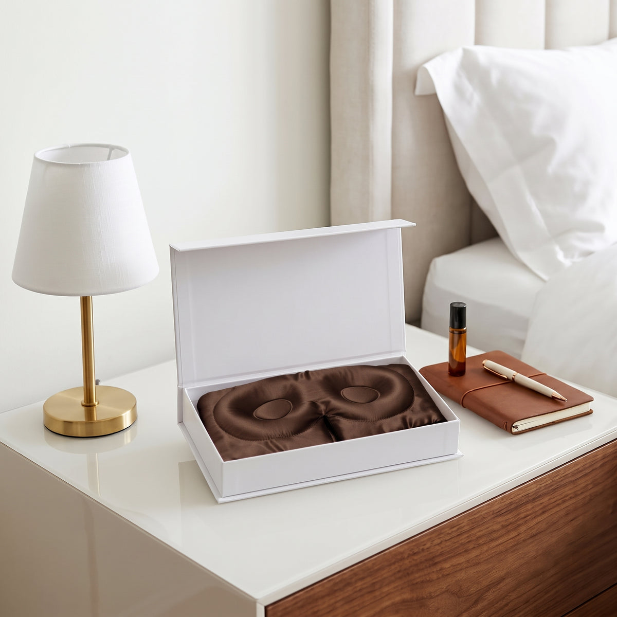 A white gift box is open on a bedside table, revealing a dark brown eye-shaped massage or facial therapy tool inside. The scene is styled like a bedroom with a white lamp, bed, and notebook nearby.