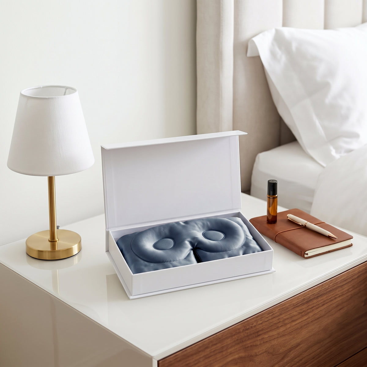 A white gift box is open on a bedside table, revealing a blue silicone massage or wellness device. A small amber bottle, a brown notebook with a pen, and a white table lamp are arranged nearby, with a bed in the background.