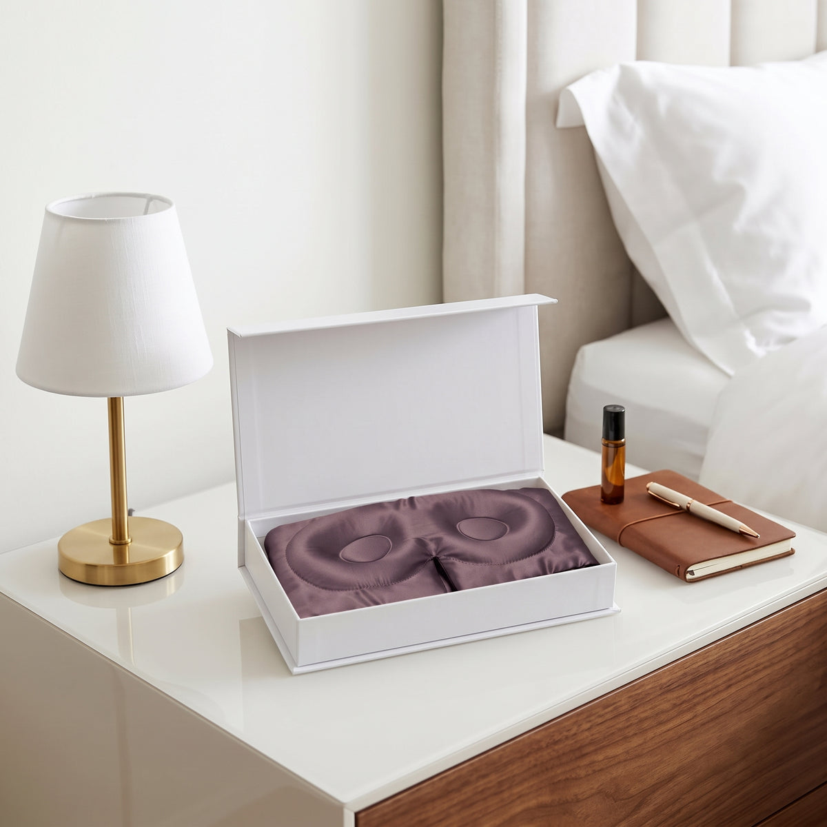 A purple eye mask set inside an open white gift box on a bedside table, styled with a gold lamp, white bedding, and a brown notebook.