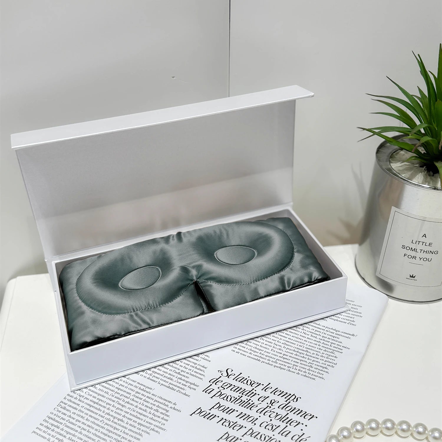 Silk eye masks in a white box on a magazine with a plant and candle in the background