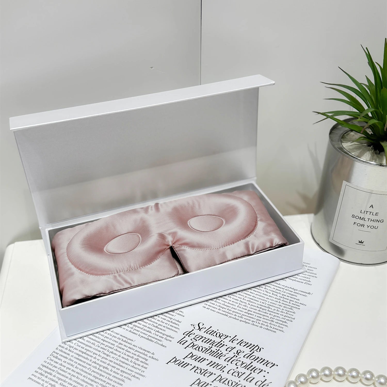 Pink sleep mask in an open white box on a magazine with a plant and candle in the background.