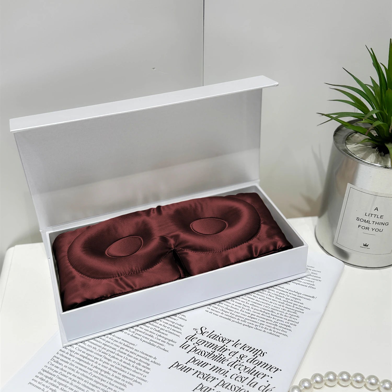 Maroon satin eye mask in a white box on a magazine with a plant and candle in the background.