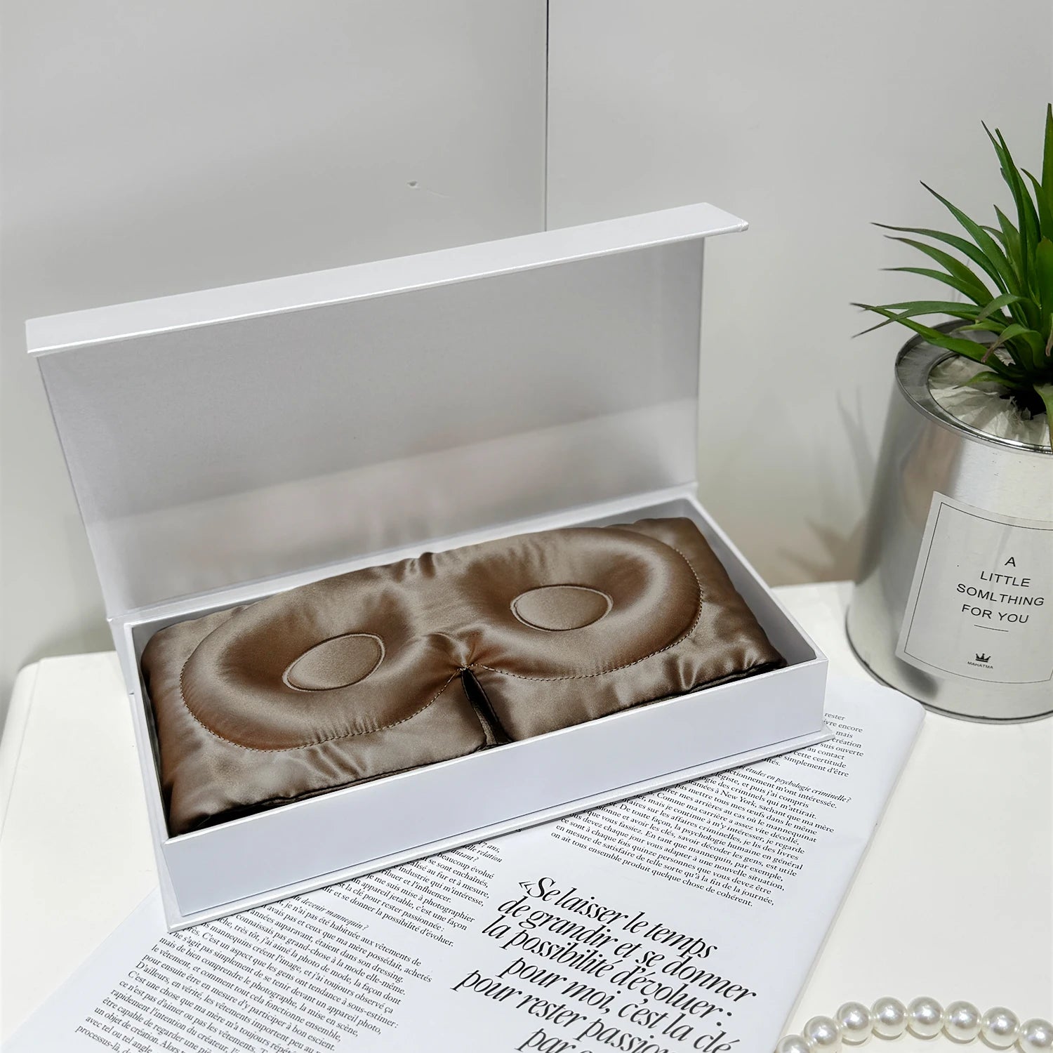 Brown satin eye mask in an open white box on a white surface with a plant and candle in the background.