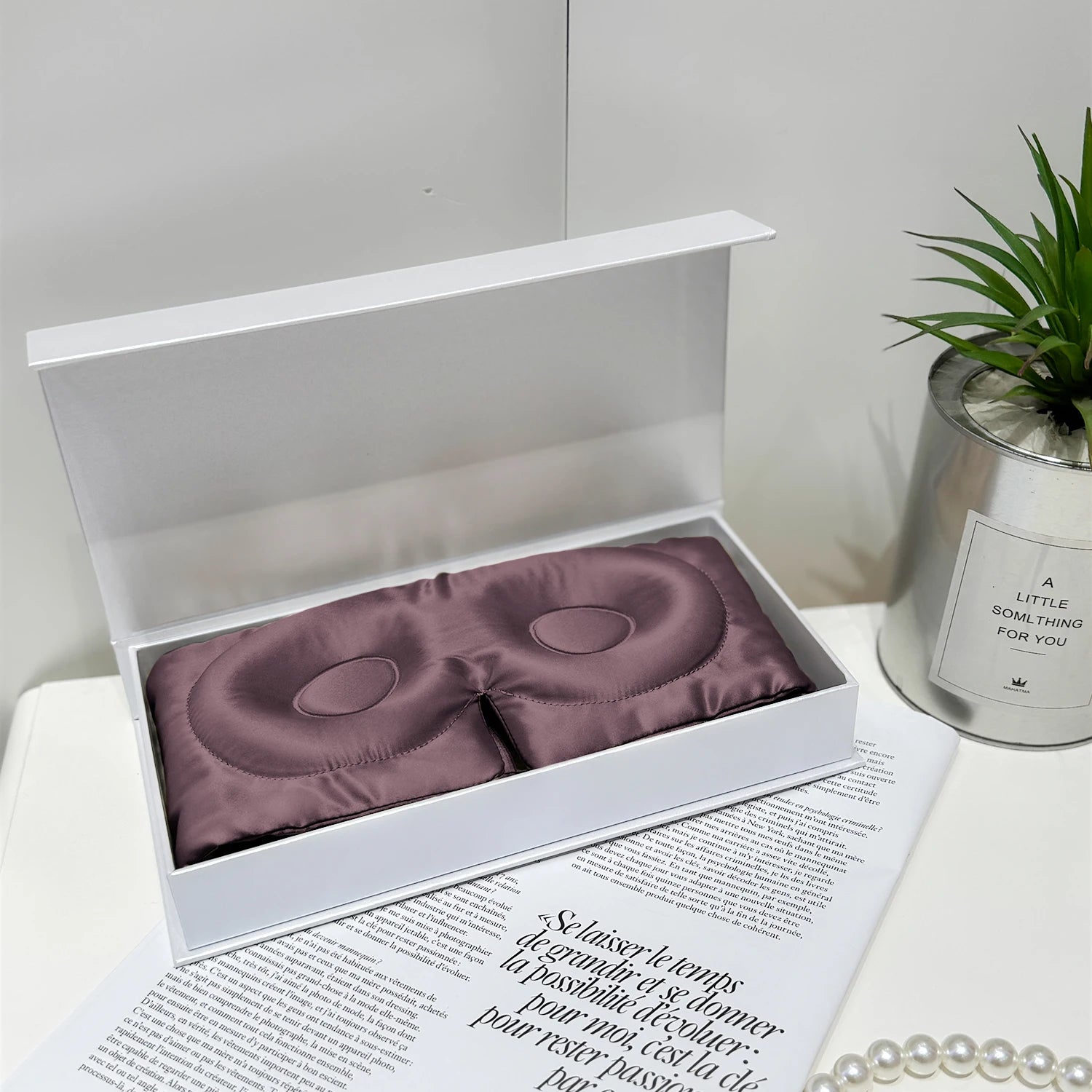 Purple satin eye masks in a white box on top of a magazine with a plant and decorative item in the background.
