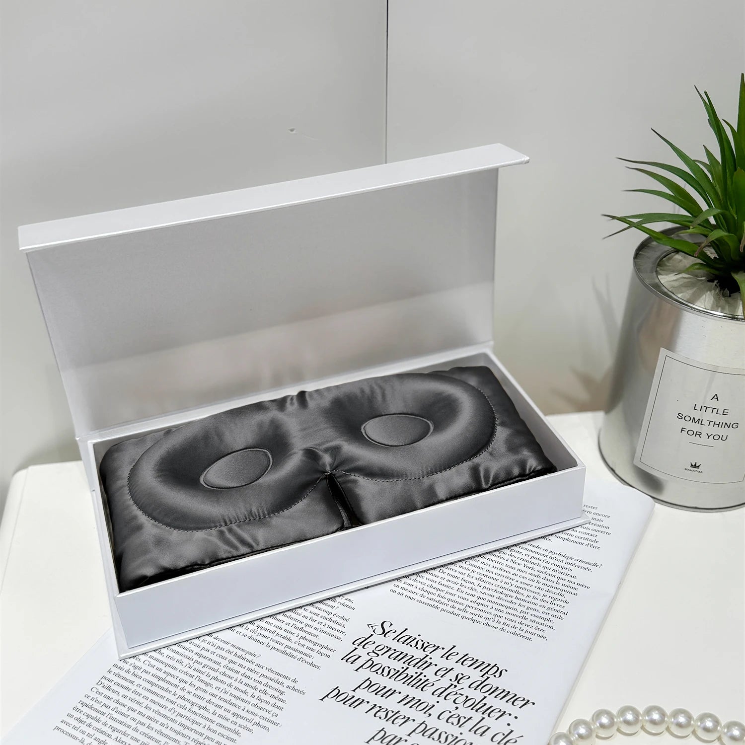 Gray eye masks in a white box on a magazine with a plant and candle in the background