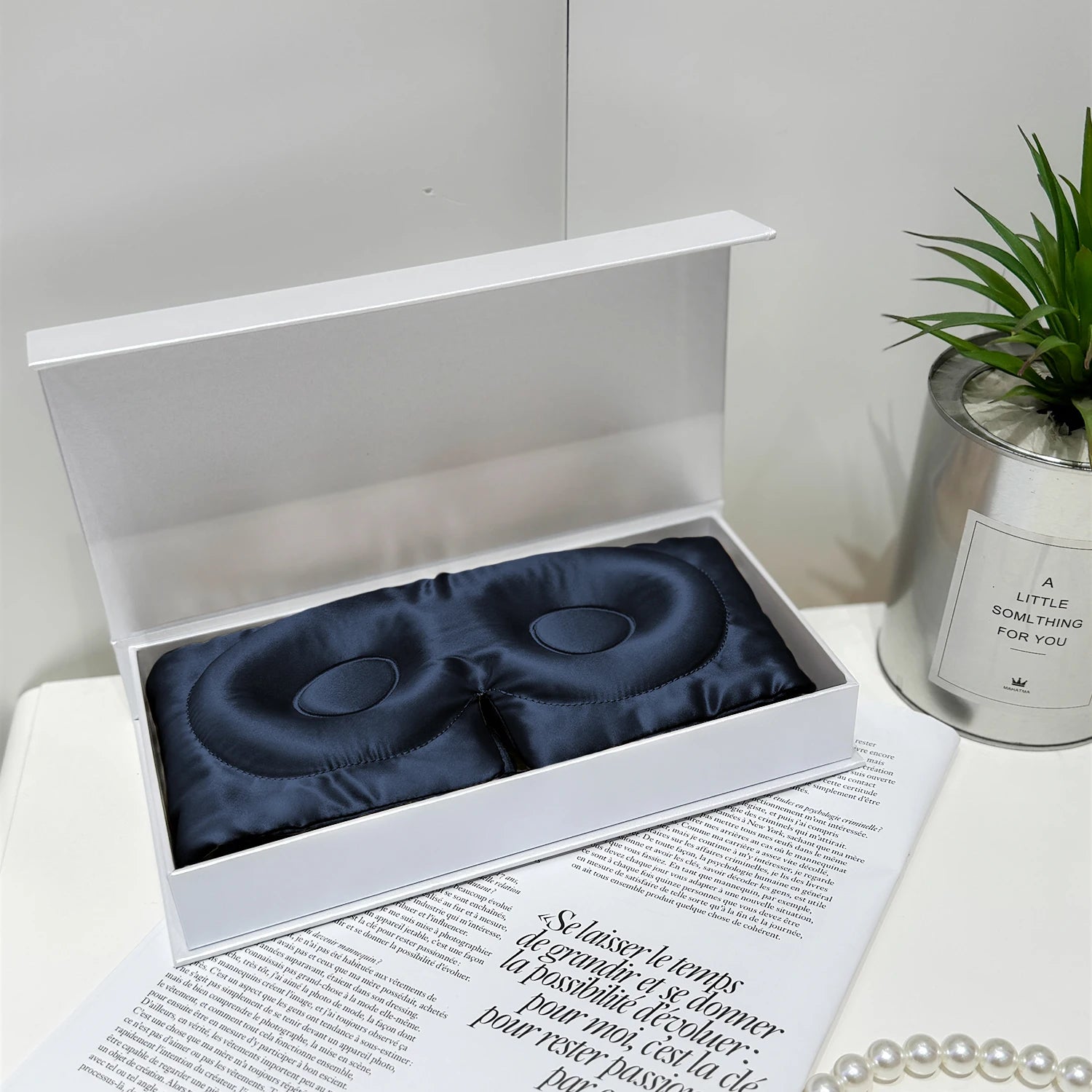 Navy blue eye masks in a white box on a magazine with a plant and candle in the background.